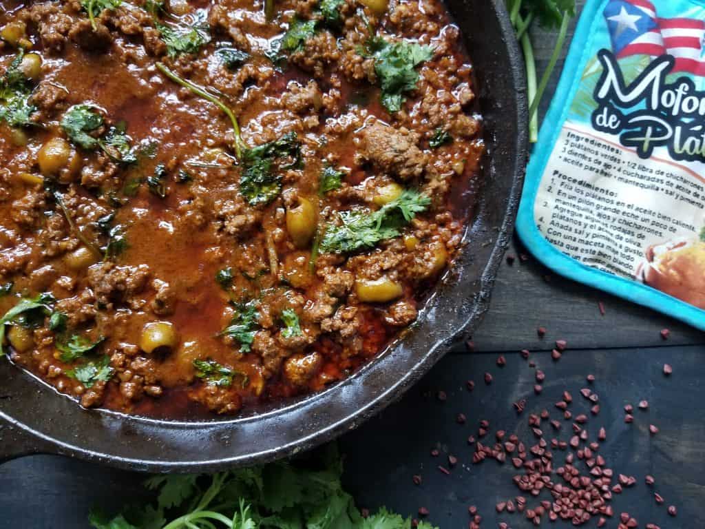 The finished Puerto Rican Picadillo in a black cast iron skillet on a wooden background. There is a blue potholder and annotto seeds on the background.