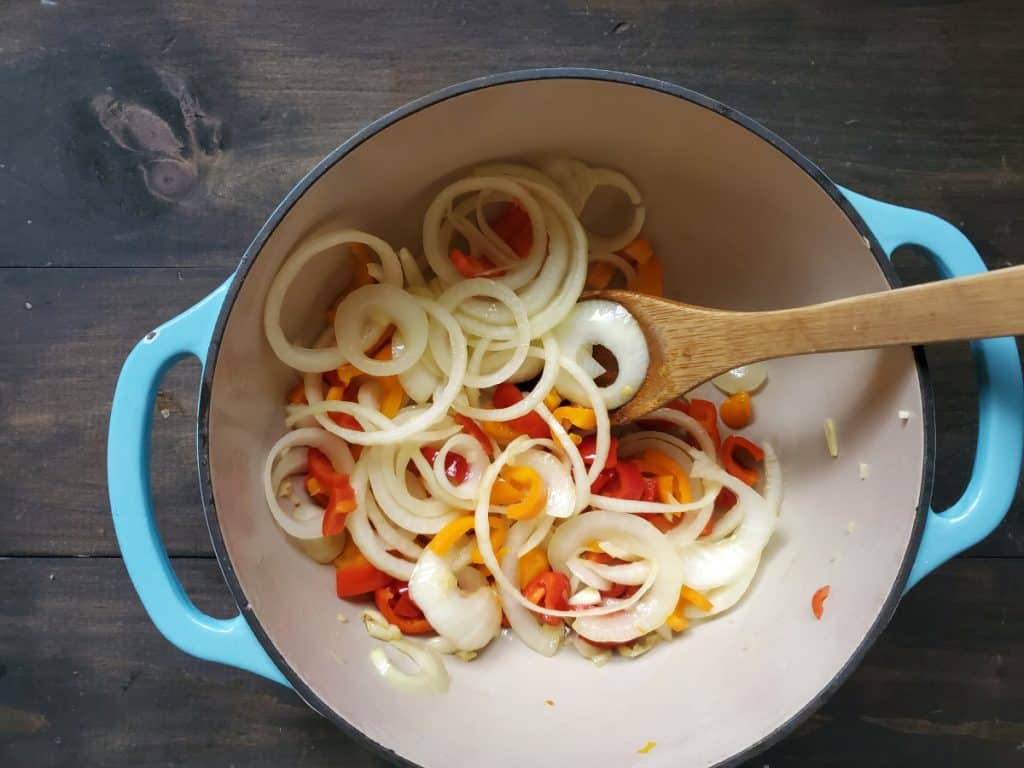 Adding the peppers, onion, and garlic to the pan and stirring it with a wooden spoon.