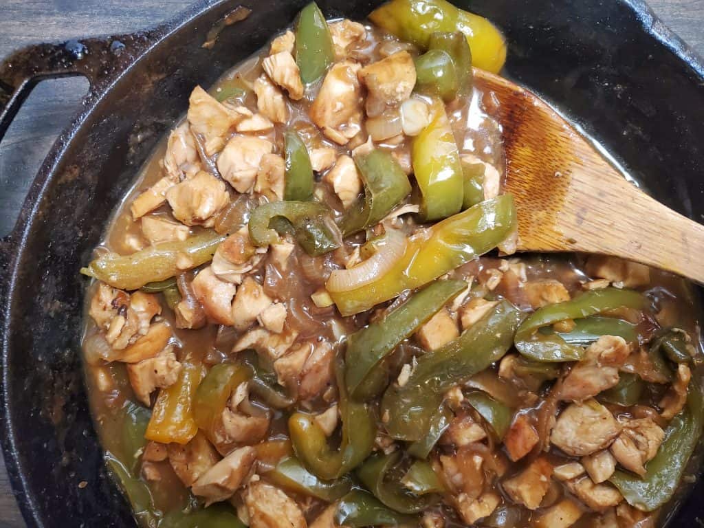 Chicken teryiyaki mixed together in a black cast iron skillet with a wooden spoon inside.
