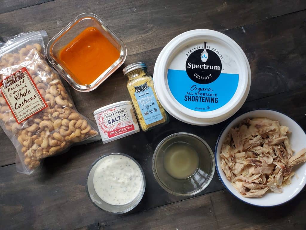 Ingredients for dairy free buffalo chicken dip are all spread out on a dark wooden background. From left to right there are cashews, buffalo sauce, ranch dressing, salt, nutritional yeast, apple cider vinegar and lemon juice mices, vegetable shortening, and chicken.
