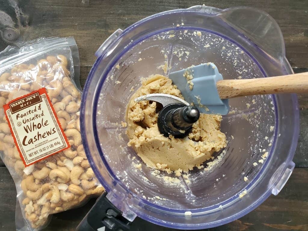 A closeup of the blended dairy free cream cheese ingredients, with a rubber spatula inside the blender. A bag of roasted and unsalted whole cashews is in the background.