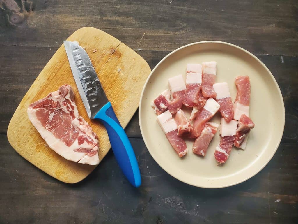 A pork chop and a knife on a cutting board. Next to it is a plate with pork cut into strips. This pork has a particularly thick rim of fat.