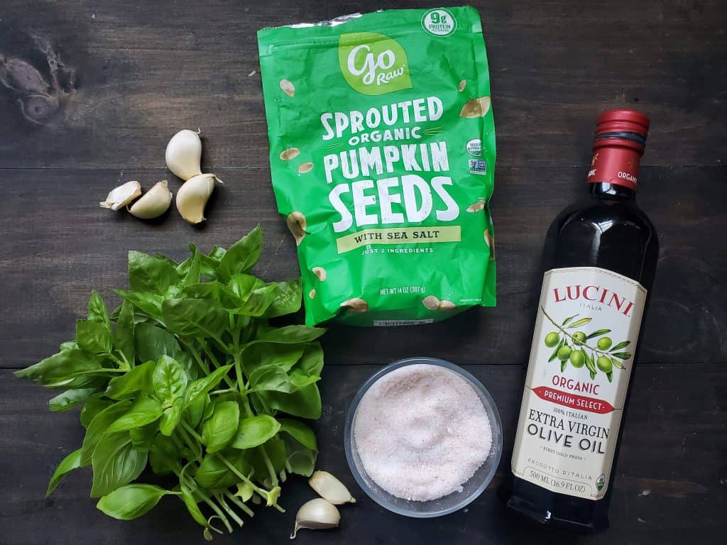 Ingredients needed for nut free pesto. From left to right there are garlic bulbs, basil, a green bag of sprouted pumpkin seeds, salt, and extra virgin olive oil.