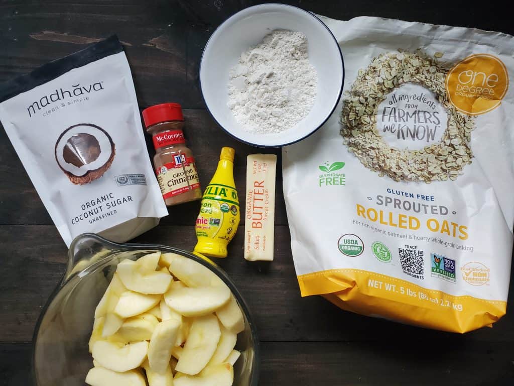 An overhead view of the ingredients in this recipe. From left to right there is organic coconut sugar, a bowl of chopped apples, cinnamon, lemon juice, butter, a bowl of flour. and a bag of sprouted rolled oats. 