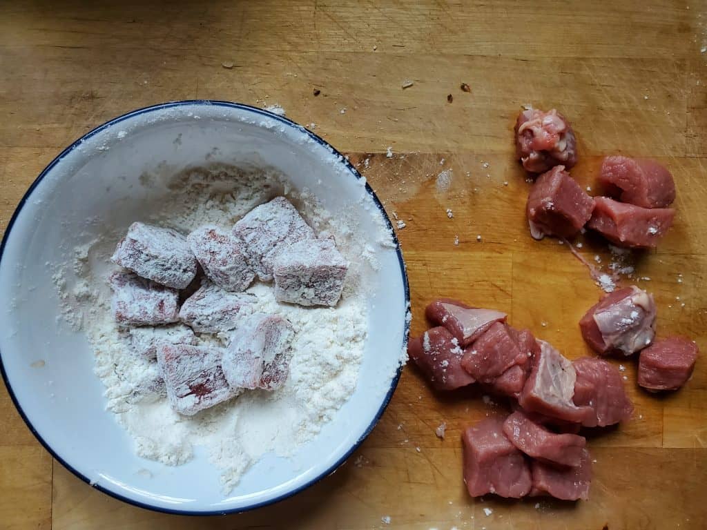 Chopped beef is on top of a cutting board, near a white bowl that contains flour with dredged pieces of meat.