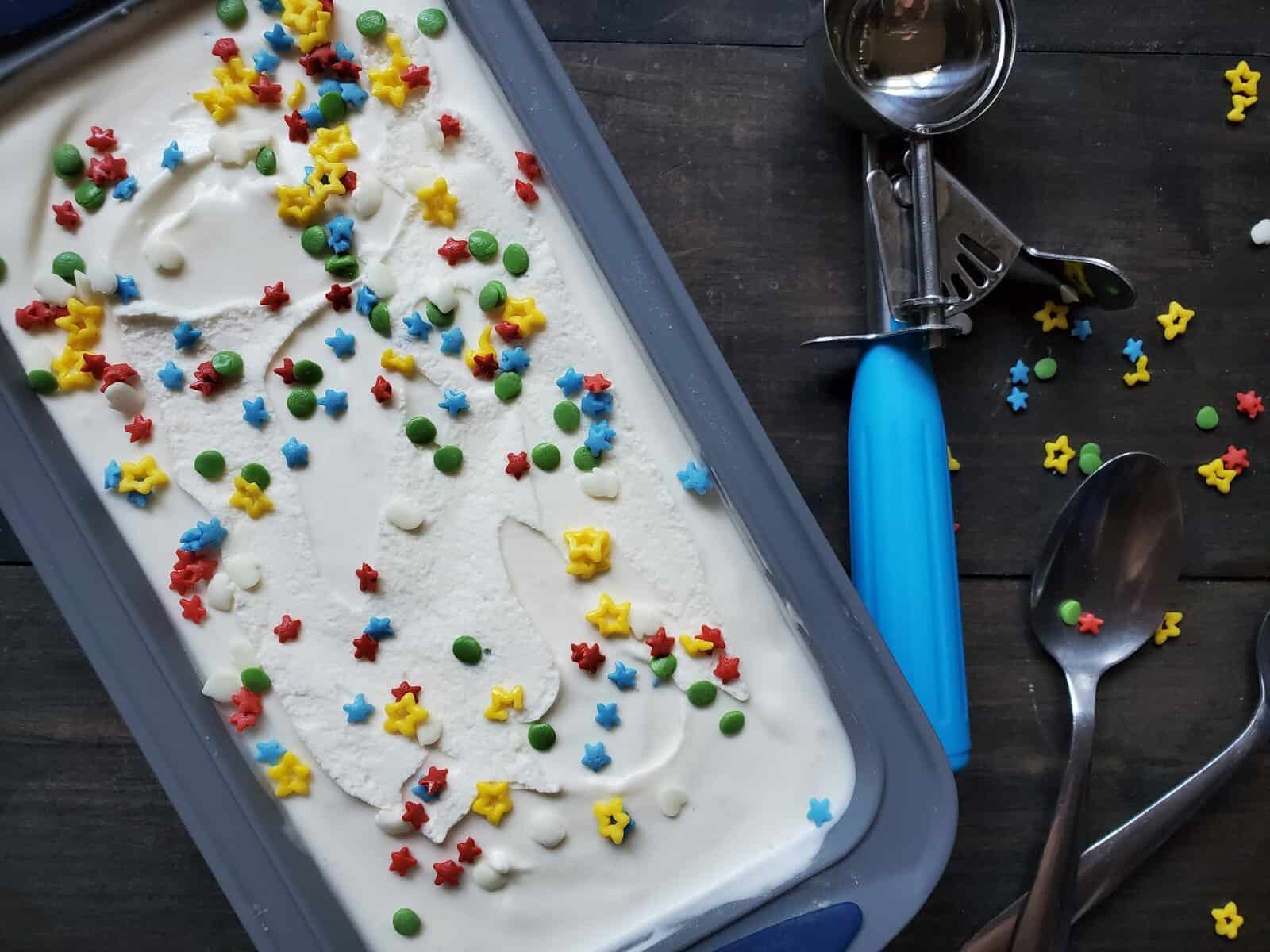 White ice cream in a container with sprinkles on top, with spoons and an ice cream scoop nearby.
