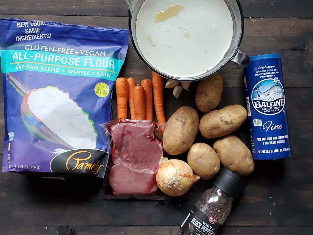 An overview shot of the ingredients needed. From left to right: Gluten free flour, meat, carrots, broth, garlic, potatoes, black pepper, and salt. Only the arrowroot powder is missing.