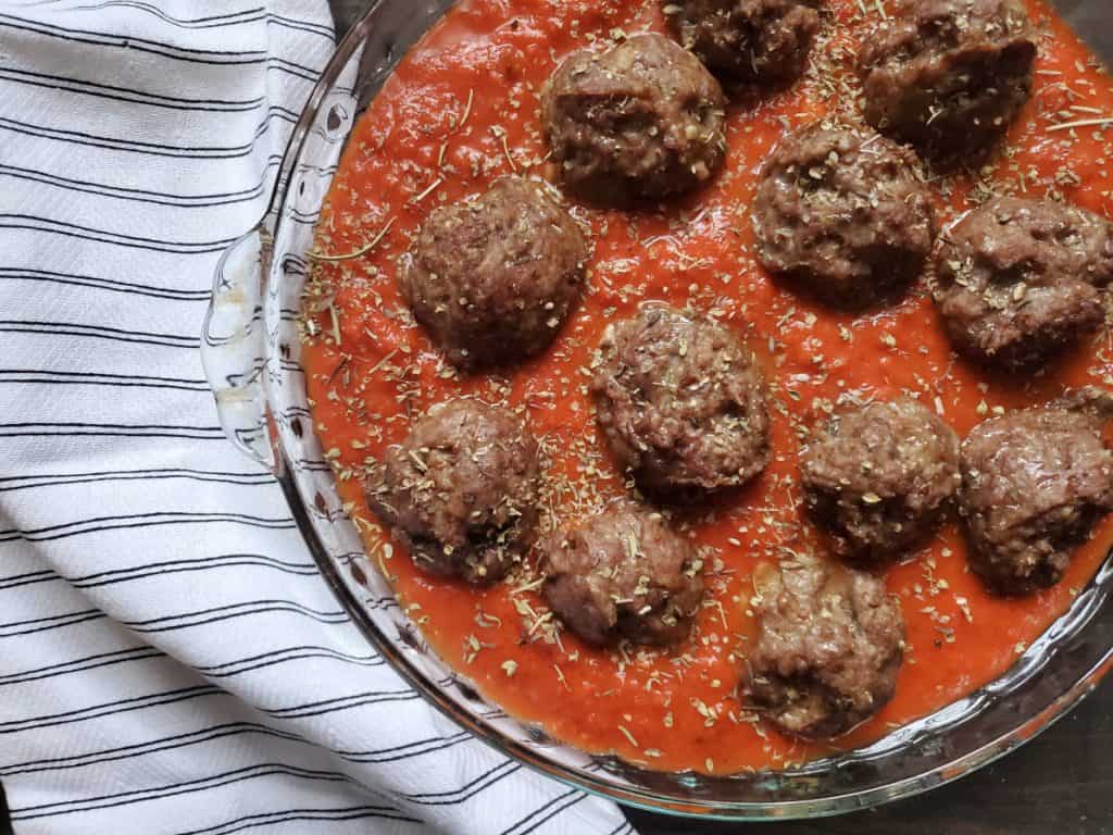 A closeup of meatballs in a bright red sauce with Italian seasoning sprinkled over the top. A black and white cloth lays next to it.
