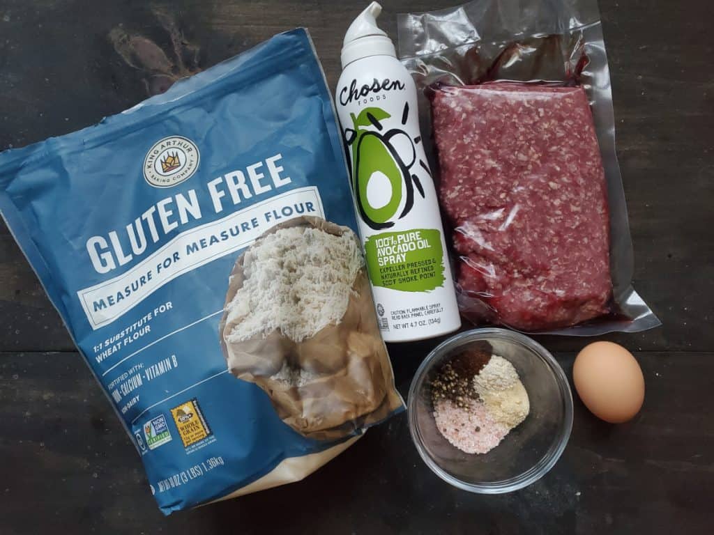 An image of the needed ingredients. From left to right, there's a blue bag of all purpose gluten free flour, cooking spray, a bowl of spices, a pack of ground beef, and an egg.