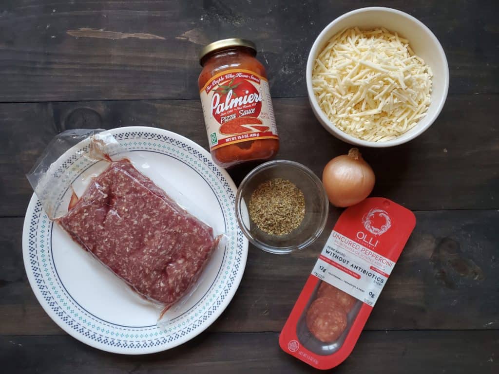 An overview of the needed ingredients. From left to right there is ground beef on a plate, a jar of pizza sauce, Italian seasoning in a bowl, pepperoni, an onion, and a bowl of cheese.