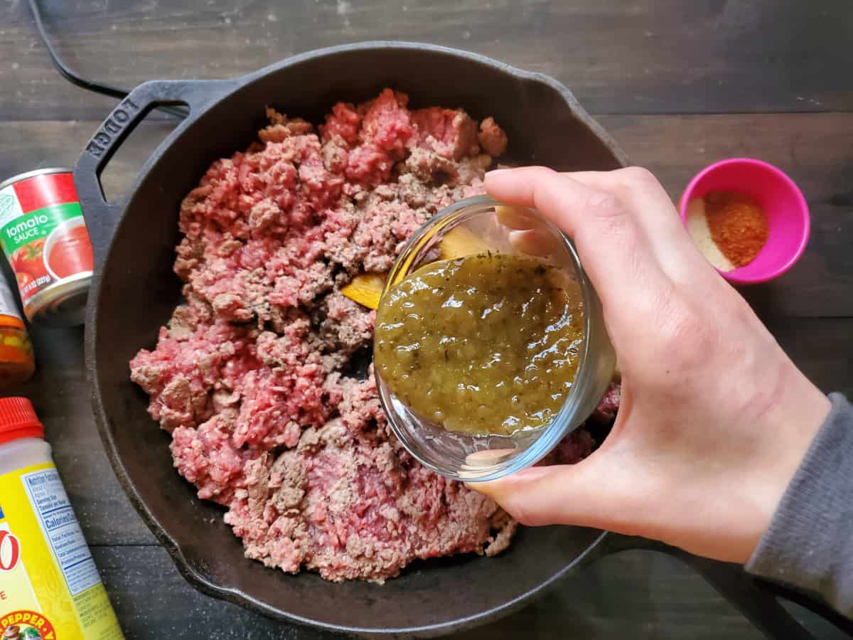 The sofrito being poured into a skillet filled with ground beef being cooked.