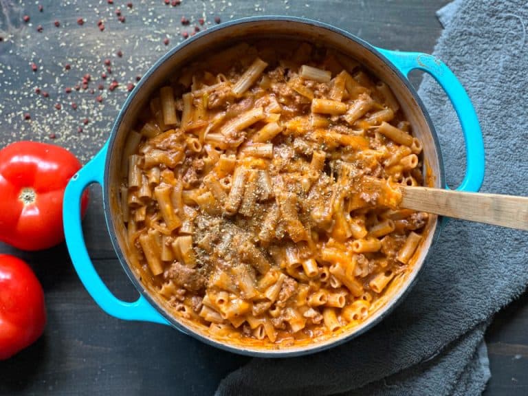 A blue pot filled with cheesy pasta mixed with ground meat, placed on a dark wooden table. A wooden spoon is in the pot. Two ripe tomatoes and pepper flakes are scattered around for garnish. A gray towel lies nearby.