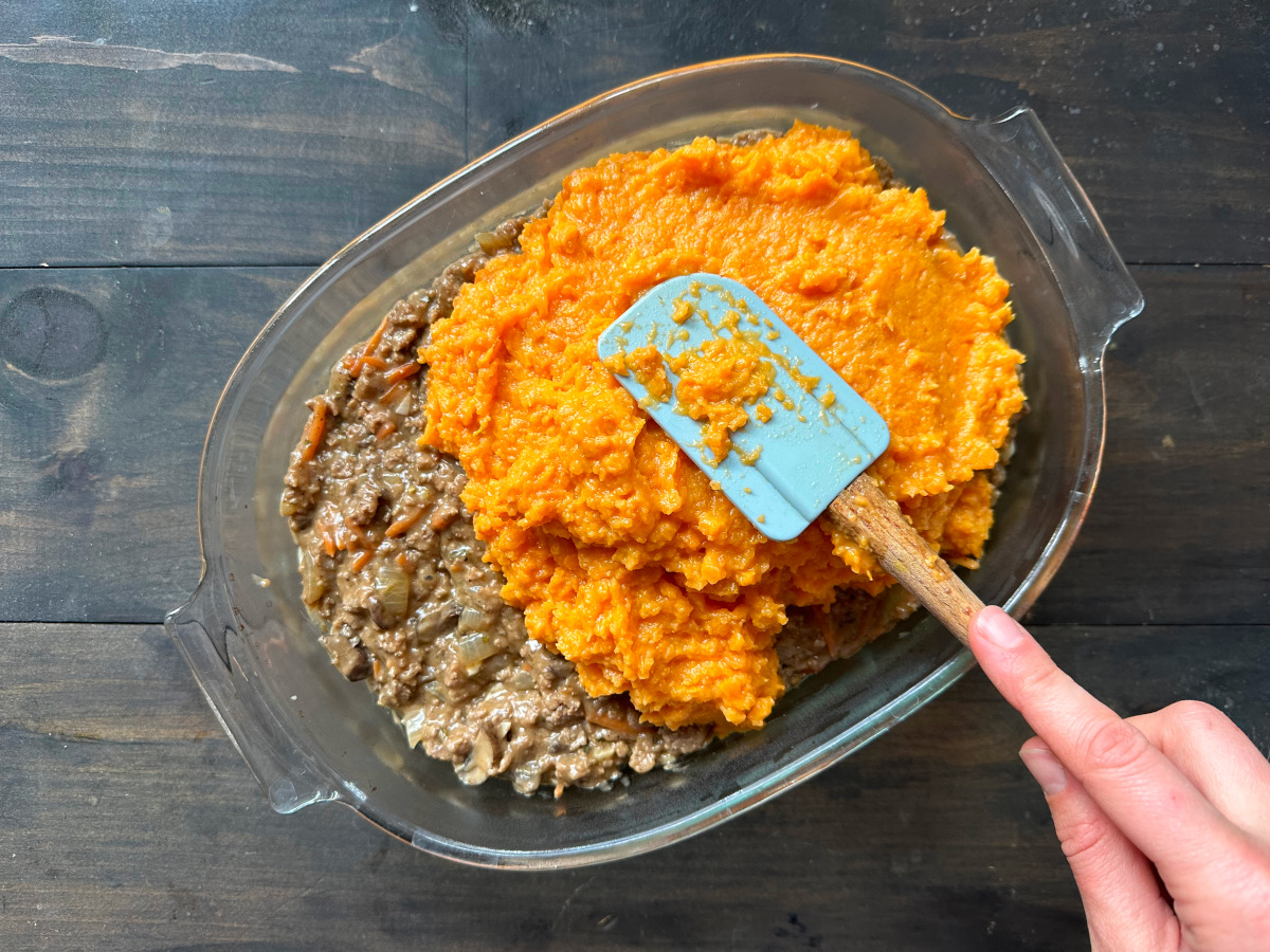 Spreading the mashed sweet potatoes over the meat mixture with a wooden spatula.