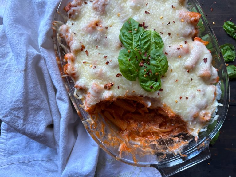 A glass dish of baked ziti topped with melted cheese and basil leaves. A portion is missing, revealing pasta in tomato sauce. A white cloth is on the side, and there are small basil leaves scattered around.
